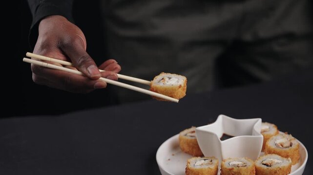 African male hand picking sushi with chopsticks over black background.