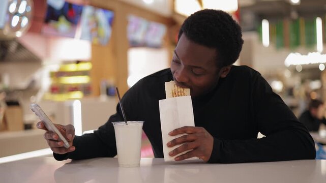 Young african male use phone and enjoying snack shaverma with chicken and milkshake at food court, fast food in mall shop.