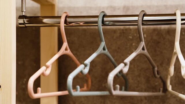 A close-up of a row of plastic coat hangers in pastel shades (beige, grey, white, peach) hanging neatly on a metal rod. Soft lighting creates a warm, minimalist atmosphere.