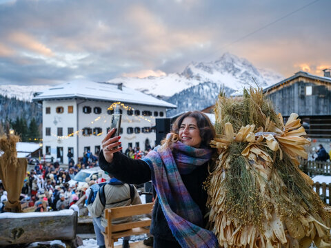 in the traditional Sauris mountain Carnival, A smiling woman takes a selfie with a phone, wearing a colorful scarf and standing next to a straw figure. Sauris,Friuli Venezia Giulia,Italy