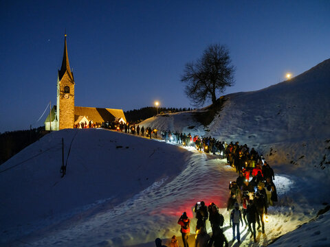 Night view of a snow-covered hill with a church,  San Lorenzo, illuminated on top and the Sauris carnival procession of people with lights walking down. Sauris,Friuli Venezia Giulia,Italy