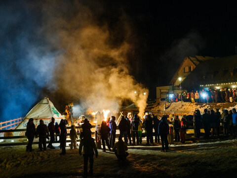 During the carnival procession, a  crowd gathers around a bonfire at night, smoke rising into the dark sky. A building is visible in the background. Sauris,Friuli Venezia Giulia,Italy
