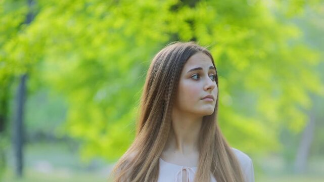 Portrait of a girl with long hair calmly observing her surroundings in a green park with a slightly alert cautious expression. Natural daylight, soft focus trees, a relaxed outdoor lifestyle context.