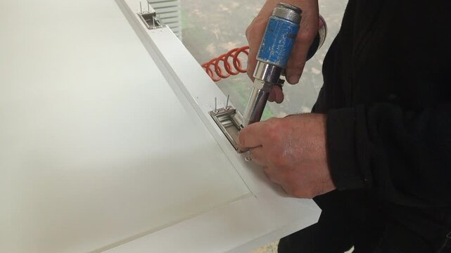 Professional worker installing metal hinges on a cabinet door using a riveting machine.