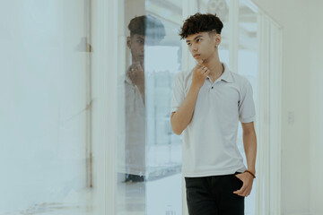 Obraz premium Handsome young asian man standing by the window, thoughtfully looking out with his hand resting on his chin. He appears pensive and deep in thought.