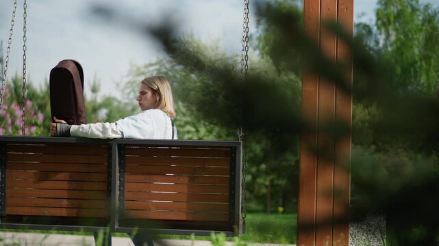 Young guitarist on wooden swing strumming near closed case under pergola in lush park, sunlight filtering through trees, contemplative songwriting session, acoustic instrument resting on lap, gentle