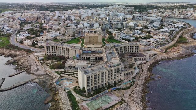 Aerial view of a derelict hotel in Malta, showing its deteriorated structure, graffiti-covered ruins, and the urban landscape of Marsaskala with the renovated Saint Thomas Tower.