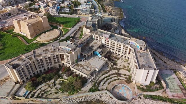 Drone view of a large abandoned coastal resort complex in southern Malta, showing deteriorating concrete structures, urban decay, and shoreline surroundings, captured along the Mediterranean coast.