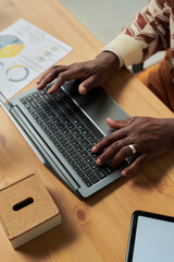 Black man typing on laptop at desk, hands with wedding ring, working on project with financial charts and tablet nearby, adult engaged in professional activity
