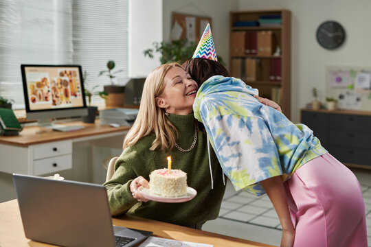 Adult woman smiling while receiving hug from young woman wearing party hat, holding birthday cake with lit candle in office setting, celebrating special occasion