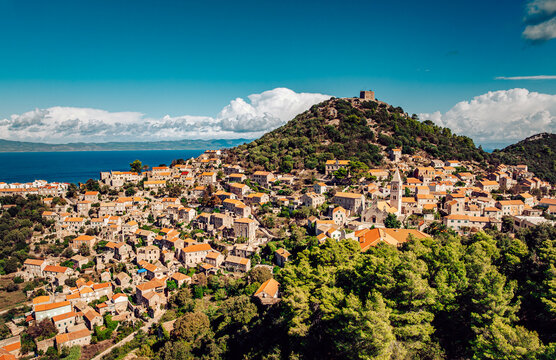 Aerial view of terracotta rooftops cascading down a verdant hillside towards the shimmering Adriatic Sea, crowned by an ancient stone fortress, Lastovo, Dubrovnik-Neretva County, Croatia.