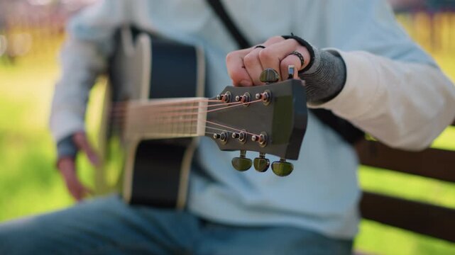guitarist tuning acoustic guitar on bench, adjusting pegs and strings, closeup of hands with ring and bracelet, casual shirt and denim, strap over shoulder, sunlight and green bokeh background,