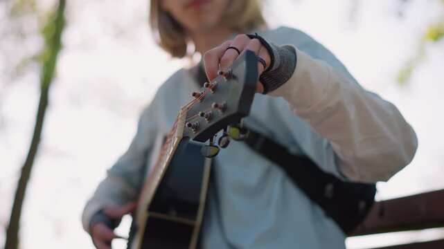 caucasian guitarist tuning acoustic guitar outdoors, busker adjusts headstock pegs, songwriter tests strings, student practices chords, denim sleeve and ring visible, bench under sunlight, soft bokeh