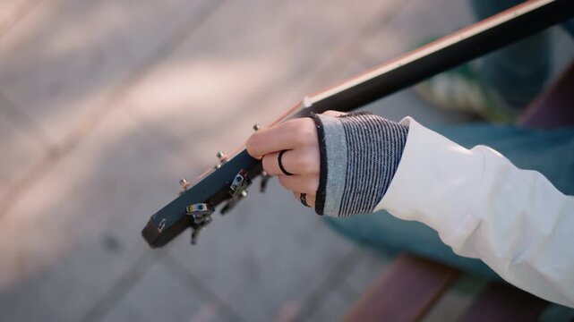closeup caucasian busker adjusting guitar tuners under soft sunlight, fingerless glove and denim visible, topdown bench angle, quick finetuning before busking, candid urban scene, upbeat lofi energy