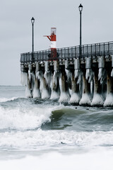 Fototapeta premium Winter pier extending over the sea, ice-covered pillars, street lamps, overcast sky, waves crashing at the shore.