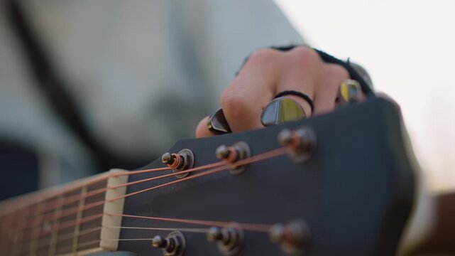 Guitarist tuning acoustic guitar headstock outdoors, hand adjusting tuning pegs, warm copper strings, wood grain, denim shirt, slow delicate motion, focused practice atmosphere, intimate musical craft