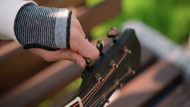 white hand tuning acoustic guitar outdoors wearing fingerless brace, closeup headstock with bronze strings, warm bokeh background, bench setting, gentle sunlight, musician practicing recovery