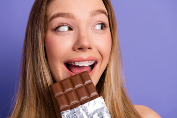 Portrait of a young woman enjoying a chocolate bar with a joyful expression on a purple background