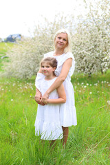 Fototapeta premium A mother embraces her daughter from behind in a blooming orchard, both in white dresses, smiling softly amid green grass and cherry blossoms, radiating love, protection, and springtime serenity.