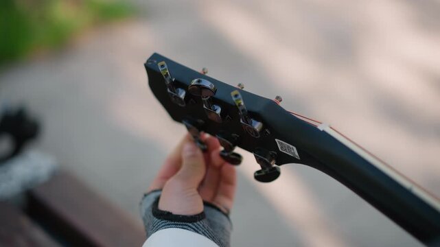 Musician adjusting guitar strings meticulously. Hands precisely tuning headstock under gentle sunlight. Focused musician carefully adjusts tuning pegs on black guitar with soft natural light