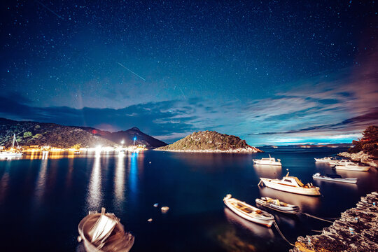 View of the bay's tranquil waters reflecting the starry sky above, illuminated by the soft glow of distant lights, contrasting with the dark silhouettes of boats and islands, Lastovo, Croatia.