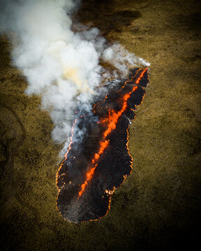 Aerial view of a long, narrow strip of vibrant orange flames cutting through a field of golden grass, smoke billowing upwards, Iceland, Iceland.
