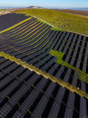 Rows of solar panels cover land in a green area under a clear sky during the day near a natural landscape with hills