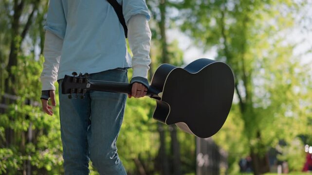 young man walking with guitar case through sunlit park, wearing white sweater and jeans, shoulder strap over chest, carrying acoustic instrument by side, relaxed stride past wooden fence and green