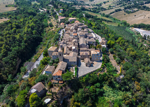 Aerial view of a medieval village atop a hill, its stone buildings and terracotta roofs contrasting with the lush green trees and distant fields, Torre di Palme, Marche, Italy.