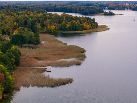 Aerial view of the serene lake bordered by reeds and vibrant autumn foliage, with a small dock extending into the calm waters, Trakai, Lithuania.