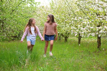 Fototapeta premium Two girls walk hand-in-hand through a sunlit orchard in full bloom, smiling, grassy path, white blossoms, embodying spring joy, sisterhood, and gentle connection with nature.