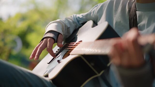 closeup acoustic guitar hands playing outdoors, busker strumming warm melody with denim sleeve and finger movement on fretboard, capo and tuning peg details, sunlight bokeh background, relaxed