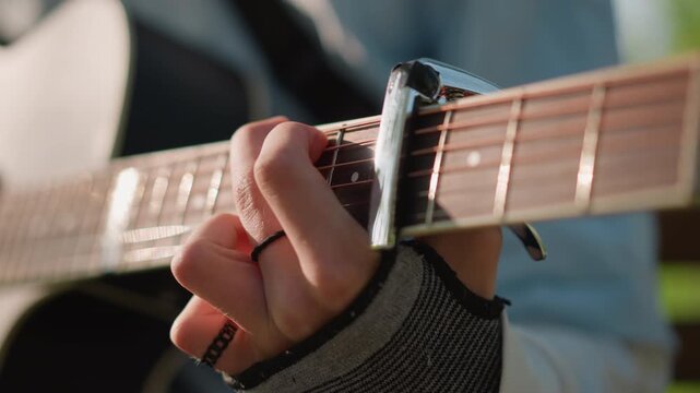closeup acoustic guitar strings capo, fingerstyle busker composing in park, warm sunlight on hands, fingerless glove and ring, detailed fretboard motion, mellow indie melody, songwriter practicing