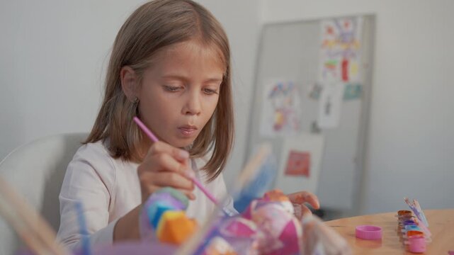 Seven year old girl sitting at table focused on her creative work painting with brushes and gouache paints expressing her creativity through art and concentration