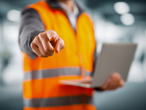 Person in a bright orange reflective safety vest holding a laptop and pointing directly at the camera with a blurred industrial background for workplace safety conce