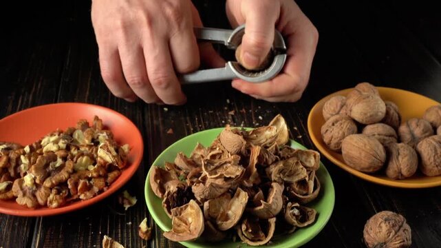 Hands cracking walnuts with a nutcracker, revealing the nut inside, surrounded by empty shells and whole walnuts on colorful plates