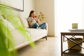 Full length view of a woman enjoying a quiet morning, relaxing on couch in bright living room, holding a warm beverage