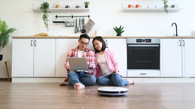 An Asian couple relaxes in their kitchen while a robot vacuum cleans the floor