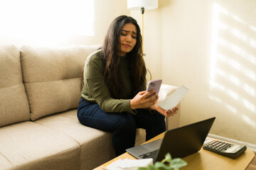 Worried young woman sitting on sofa, holding a phone and paperwork, calculating expenses and facing...