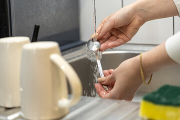 Female hands washing a metal spoon under running water in a modern kitchen sink, depicting essential daily household chores and maintaining cleanliness