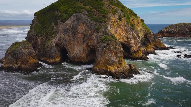 Aerial drone view of a grass-covered sea stack with a sea cave on Chilo&eacute; Island, Patagonia&rsquo;s Pacific coast, Chile. Powerful waves hit a remote sandy beach and rugged green cliffs under an overcast sky