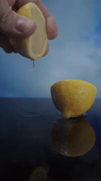 Vertical 9:16 slow motion video of a hand squeezing half a lemon as juice droplets fall onto a wet surface, while clouds move softly in the background