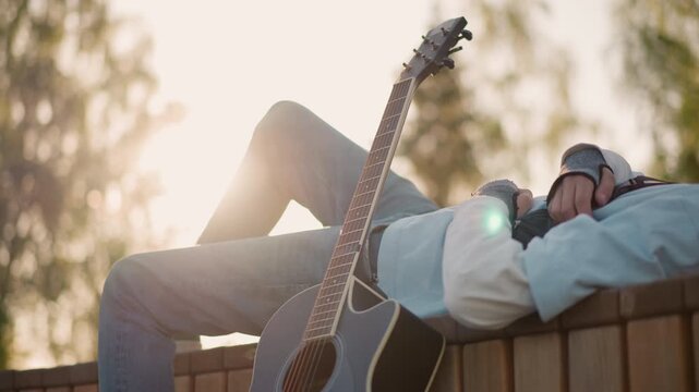 closeup acoustic guitar relaxed musician, hands folded across chest, head tilted back, leg bent over bench edge, sun flare through trees, fretboard and strings in foreground, peaceful contemplation,