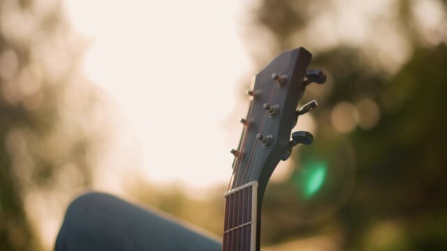 acoustic guitar headstock against sunset glow, indie guitarist tuning instrument while resting with denim knee warm backlight and lens flare create soft bokeh, intimate unplugged mood with park
