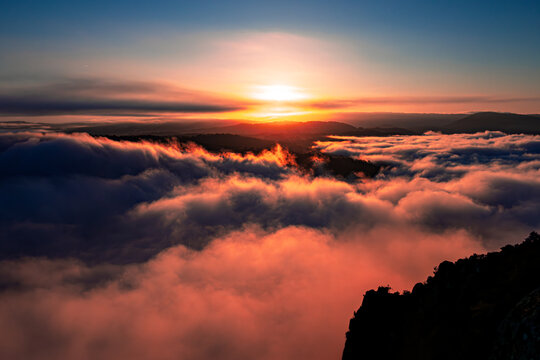 Aerial view of the sun dipping below the horizon, casting a warm orange glow over the thick blanket of clouds nestled above the Canyon du Sil, Sober, Lugo, Spain.