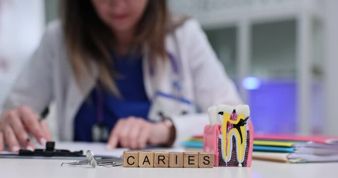 Wooden cubes spell word Caries near model tooth on dental desk. Female dentist studies chart while writing treatment plan for patient decayed tooth