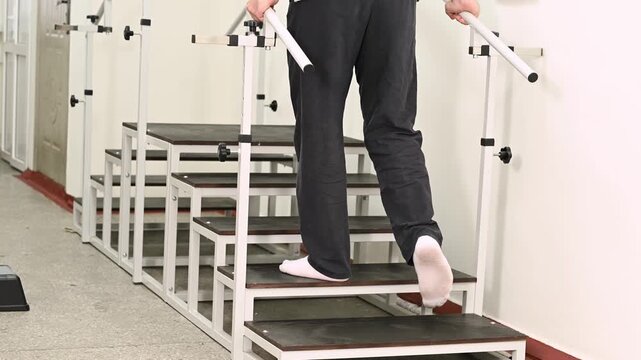 A man undergoes rehabilitation after an injury on a stair climber with parallel bars at a clinic under the supervision of a physical therapist. Close up legs on stairs