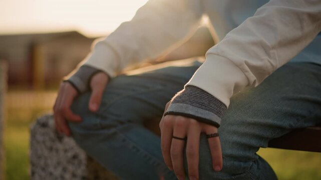 Fingers fidgeting softly beneath warm light. Calm hands resting as evening shadows gently appear. Gentle posture with hands resting on knee amid fading sunlight and quiet anticipation