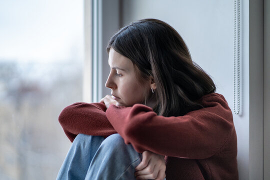 Sad reflective teenage girl sits hugging knees looking to window. School stress, teen anxiety, emotional pain, solitude. Lost depressed teenager feels inner conflict, adolescence crisis alone at home.