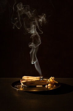 Bois de Palo Santo br&ucirc;lant sur un plateau dor&eacute;, fum&eacute;e verticale sur un fond brun fonc&eacute;